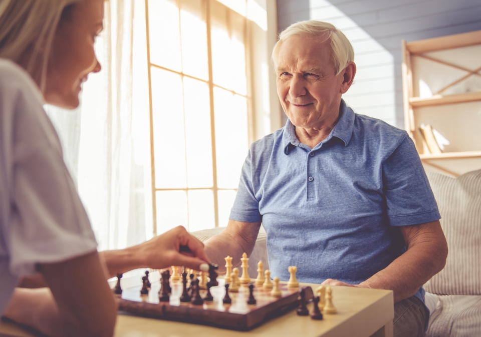 An elderly man and a younger woman play chess together in a sunlit room, showcasing elegant living space. The elderly man smiles attentively while the woman makes a move on the chessboard, their surroundings enhanced by thoughtful interior design.