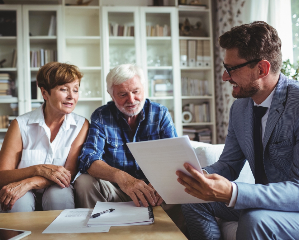 An older couple sits on a couch, reviewing documents with a man in a suit holding papers and a pen. They're new to Medicare and are discussing their enrollment options. Shelves filled with books in the background add a scholarly touch to the scene.