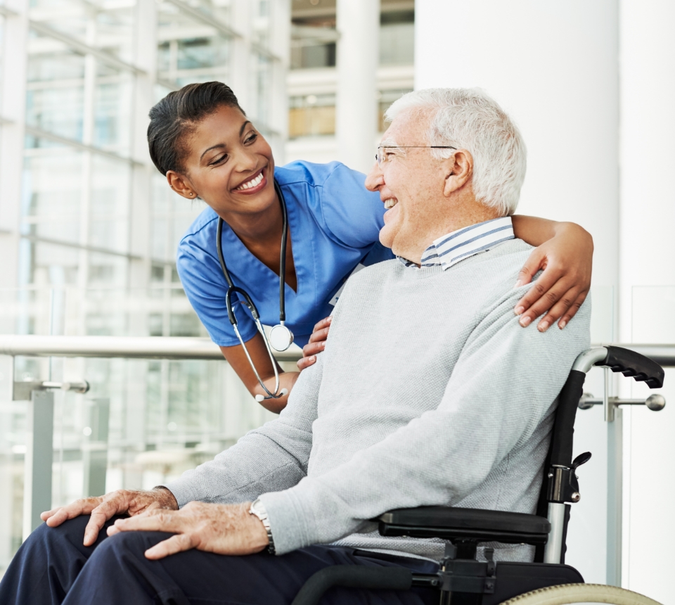 A nurse in blue scrubs smiles and embraces a senior man in a wheelchair, embodying the compassionate care core to Medicare. They appear to be in a modern medical facility.