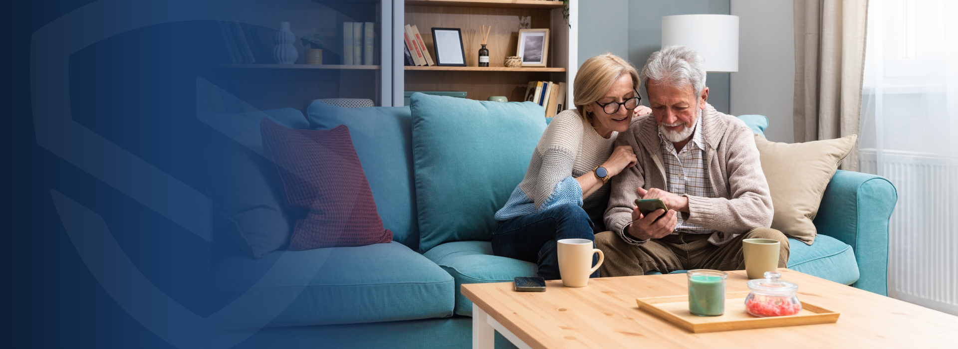 An elderly couple sits on a light blue sofa in their cozy home, looking at a smartphone together. A coffee table with a tray, two mugs, and a jar is in front of them. Shelves and a lamp add warmth to the background.