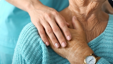 A caregiver rests their hand on the shoulder of an elderly person wearing a light blue sweater, a wristwatch, and reflecting on the benefits of Medicare.