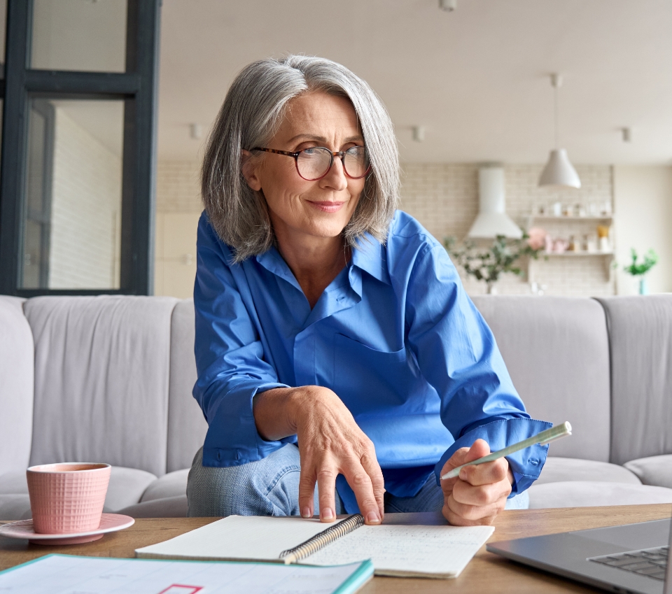 An older woman with gray hair and glasses, wearing a blue shirt, sits on a sofa writing in a notebook with a pen. A pink cup, open book, and papers are scattered on the coffee table in front of her. She appears focused, perhaps jotting down notes as she navigates her new to Medicare information.