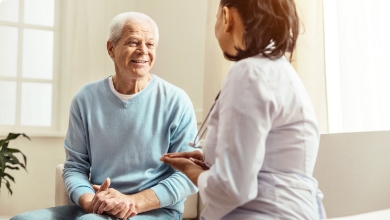 An elderly man sitting and smiling talks to a healthcare professional who is attentively listening, inside a brightly lit room with windows, discussing the importance of Medicare education.